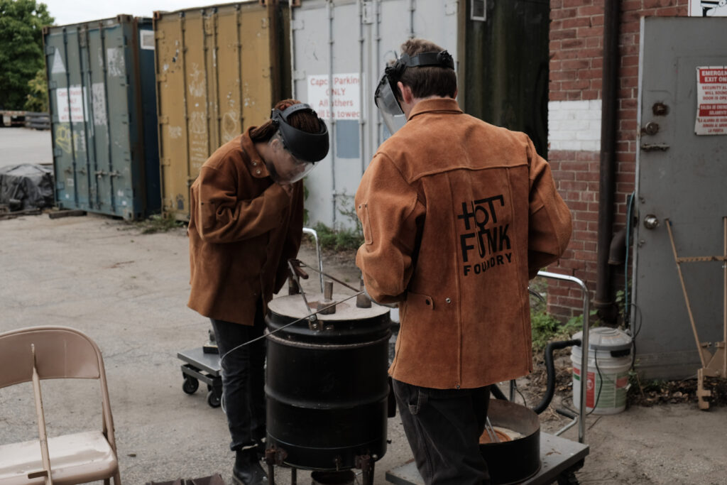 Chris Kane and Chanel (Nel) McNamara of Hot Funk Foundry at a metal pour event, wearing protective gear outdoors.