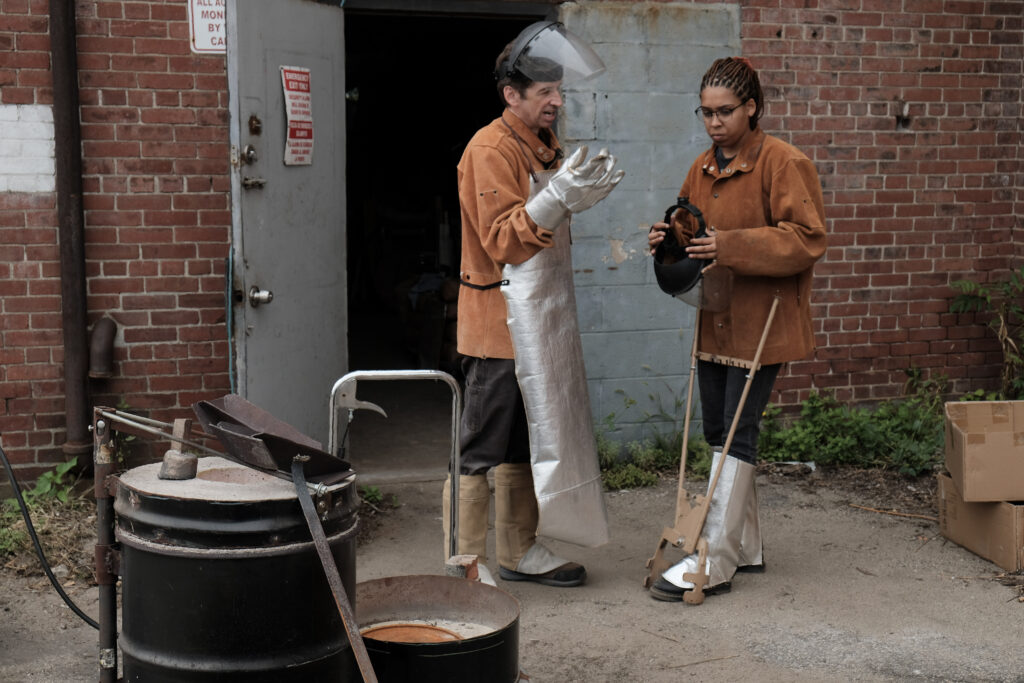 Chris Kane and Chanel (Nel) McNamara of Hot Funk Foundry discuss during a metal pour event, wearing protective gear.
