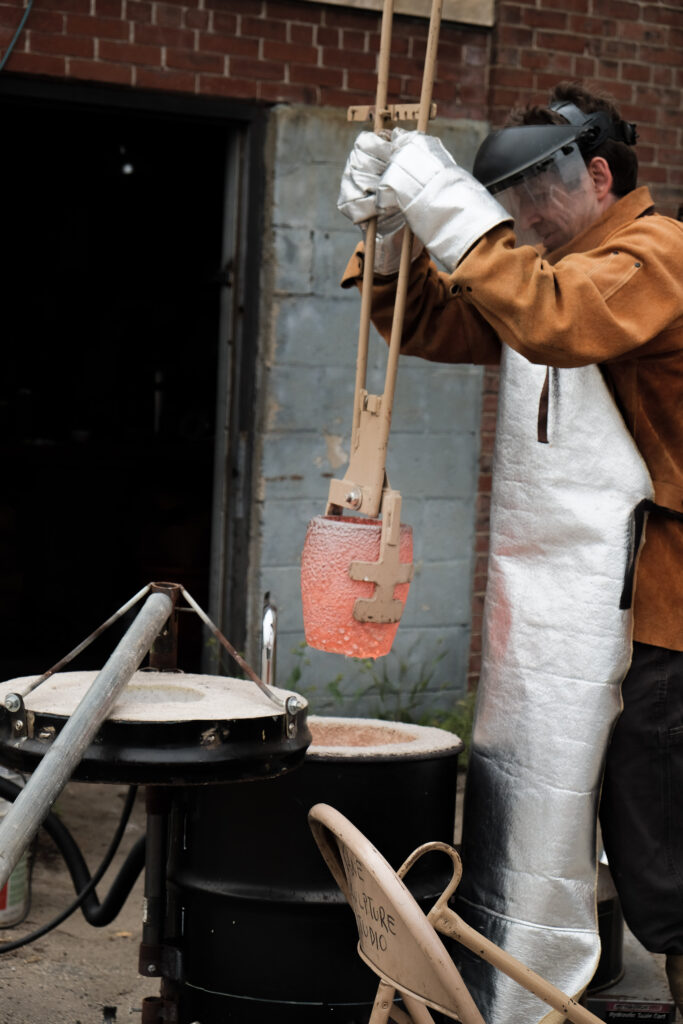 Chris Kane, founder/instructor, conducts a metal pour event, wearing protective gear and handling molten material with tongs.