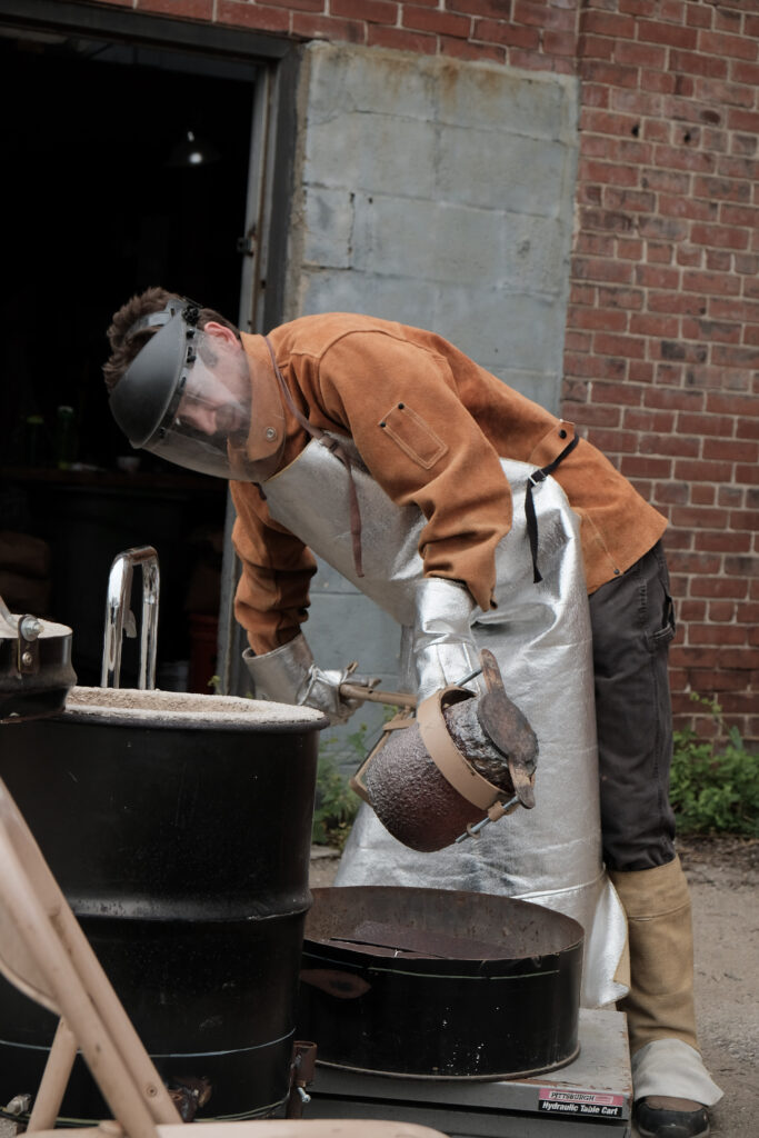 Chris Kane, founder and instructor, at a metal pour event, wearing protective gear while handling a crucible outdoors.