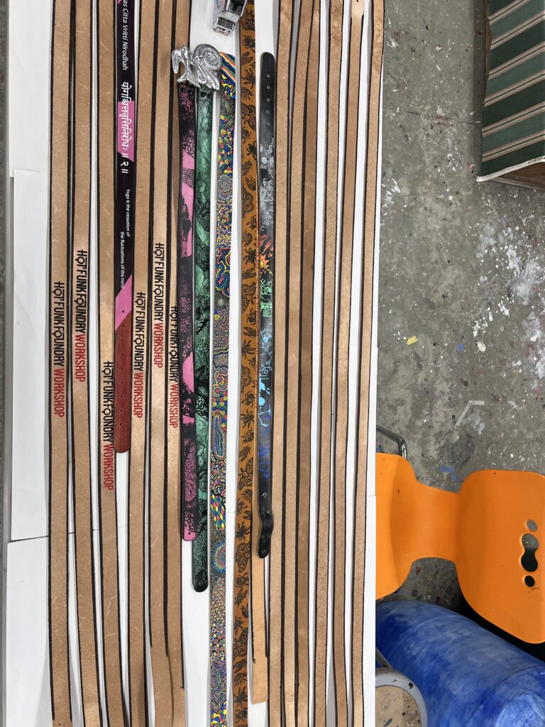 Leather straps on a table, some labeled "Hot Funk Foundry Workshop," with colorful, patterned designs and a silver buckle.