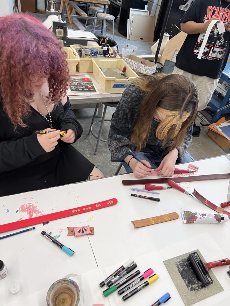 Two individuals are decorating belt buckles at the Hot Funk Foundry workshop. Art supplies and tools are scattered on the table.