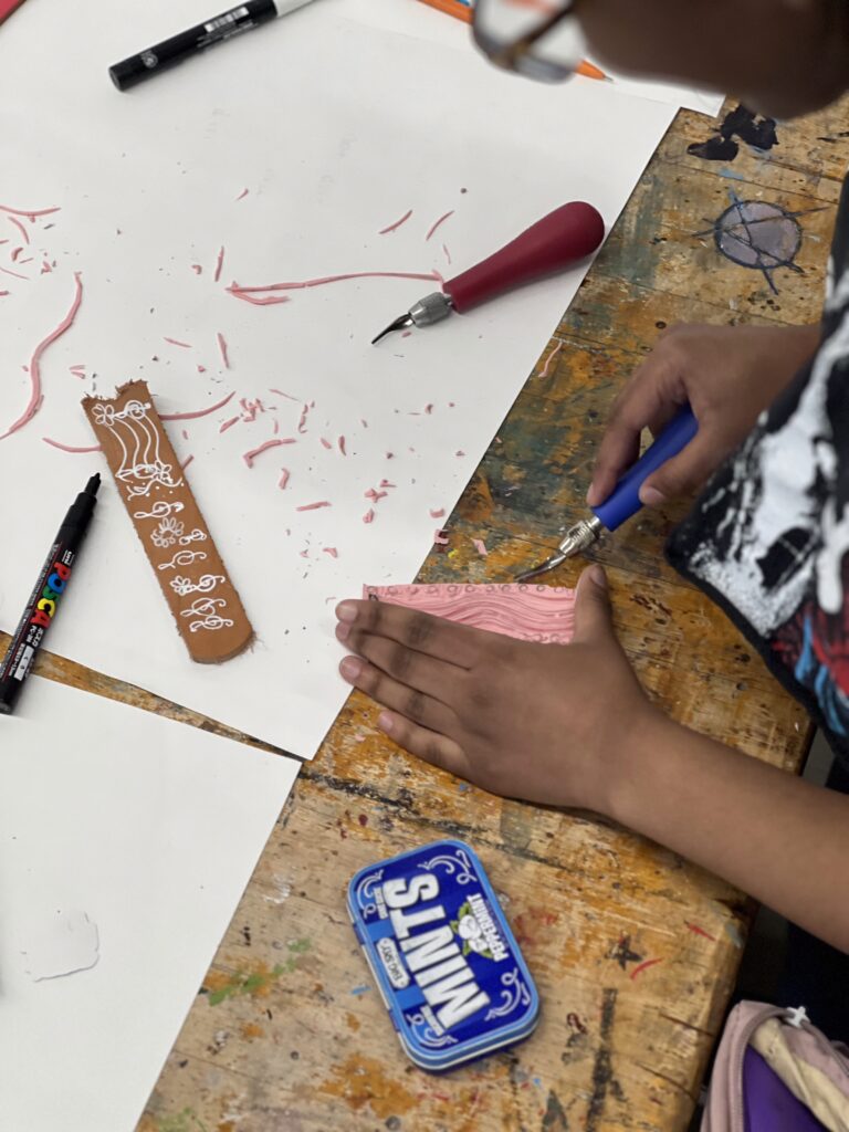 A person carves a pink design into a block during the "Hot Funk Foundry" belt buckle workshop. A mint tin and markers are nearby.