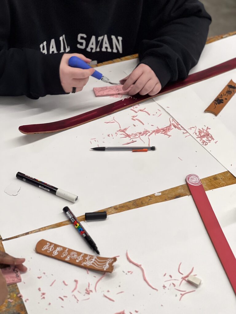 A person in a "Hail Satan" sweatshirt carves a design into a pink material at a belt buckle workshop. Markers and tools are scattered on the table.