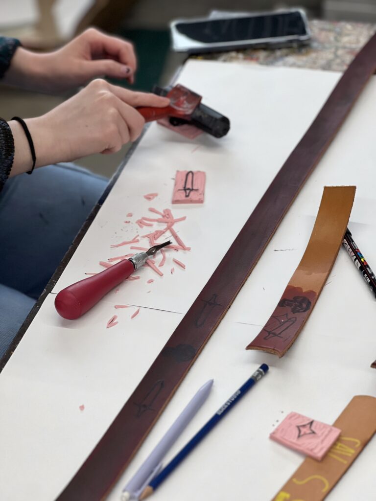 A person carves designs into pink material with a carving tool, surrounded by leather strips and art supplies on a table.