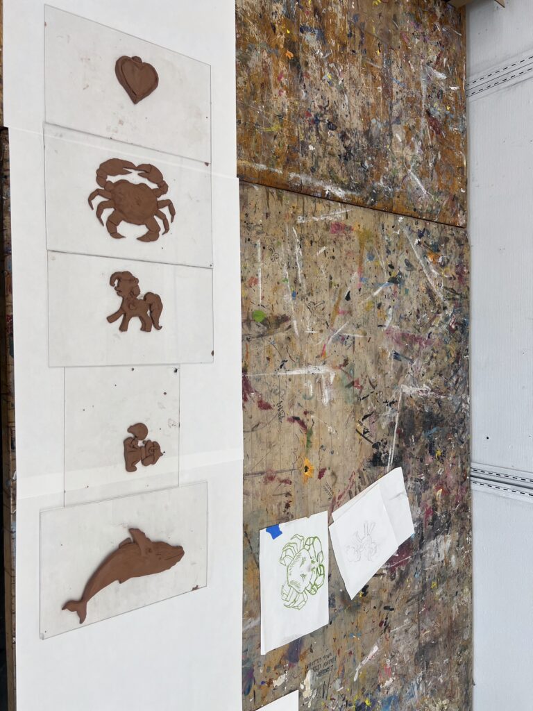 Clay molds of a heart, crab, pony, squirrel, and dolphin on white sheets against a splattered brown wall, with crab sketches nearby.