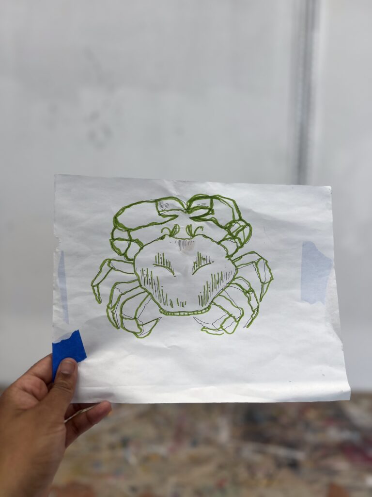 A hand holds a paper with a green line drawing of a crab, secured with blue tape, against a blurred background.