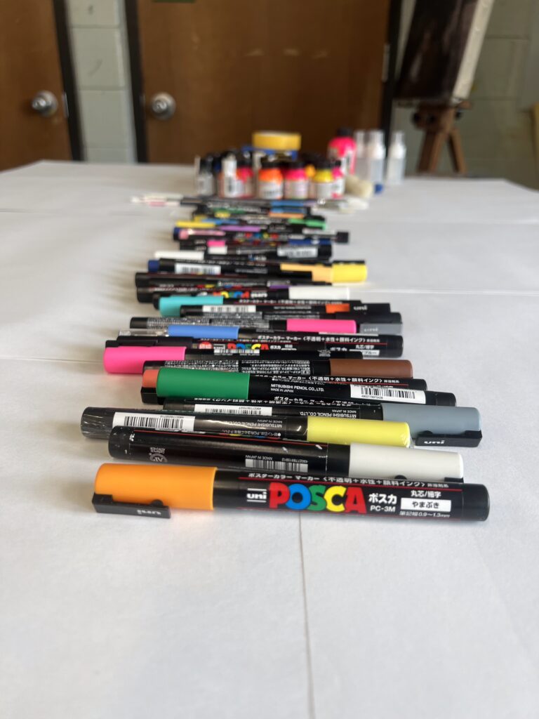 Colorful Posca markers lined up on a table with paints and art supplies in the background.