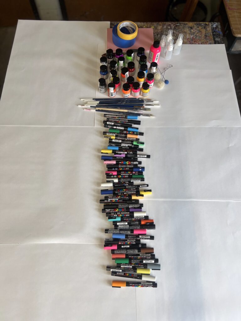 Art supplies organized on a table: paint bottles, brushes, markers, and tape on white paper.