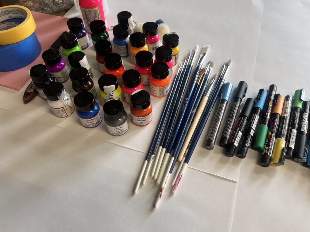 Art supplies on a table: colorful paint bottles, assorted brushes, markers, and rolls of tape arranged neatly.