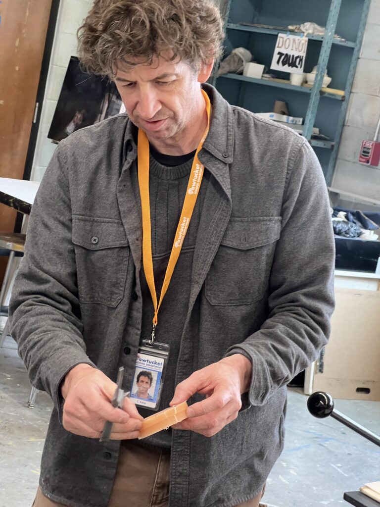 Chris Kane, the instructor, demonstrates at the "Make Your Own Cast in Bronze Belt Buckle" workshop, holding a tool and material.