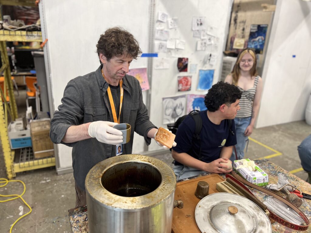 Instructor Chris Kane demonstrates casting a bronze belt buckle in a workshop, with two participants observing.