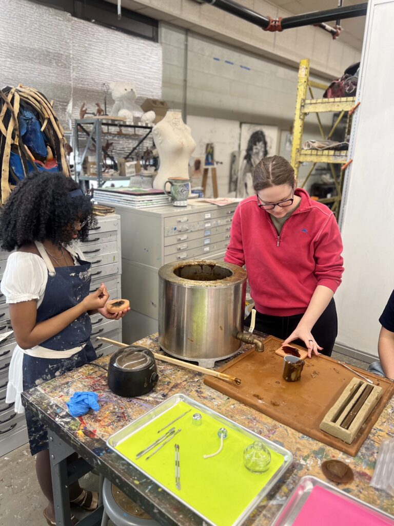 Two women in a metal art studio pour melted wax into a mold amid tools, trays, and a metal melting pot.