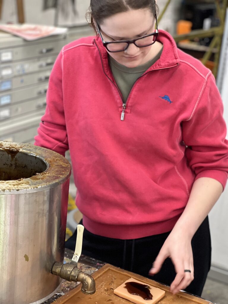 A person in a pink sweatshirt and glasses works at a workshop, pouring liquid into a mold from a metal container.