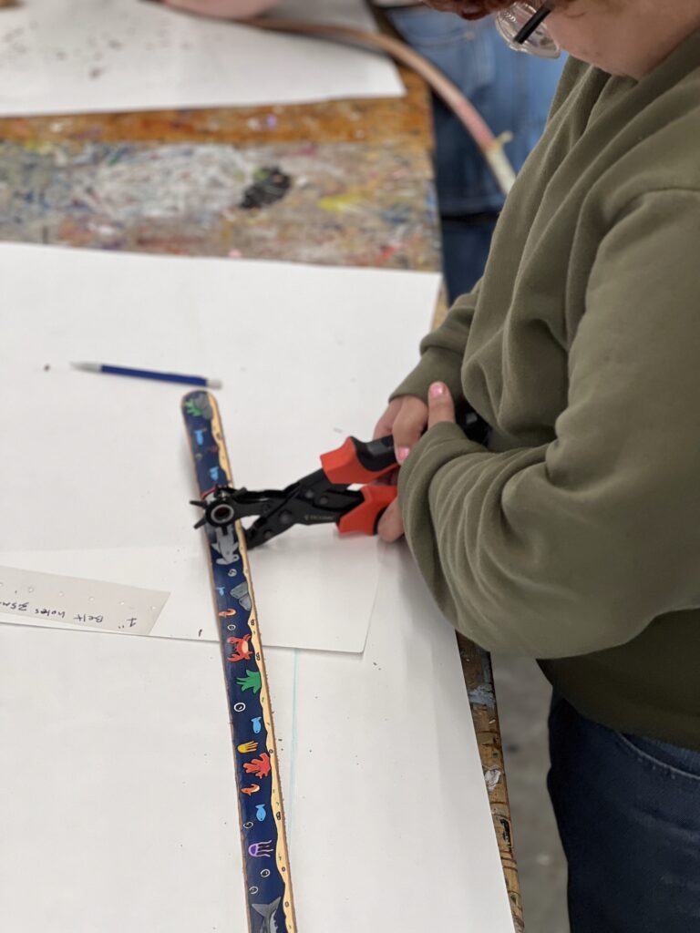 A person uses a hole punch on a colorful belt at a workshop. The table is covered with white paper and a pencil is nearby.