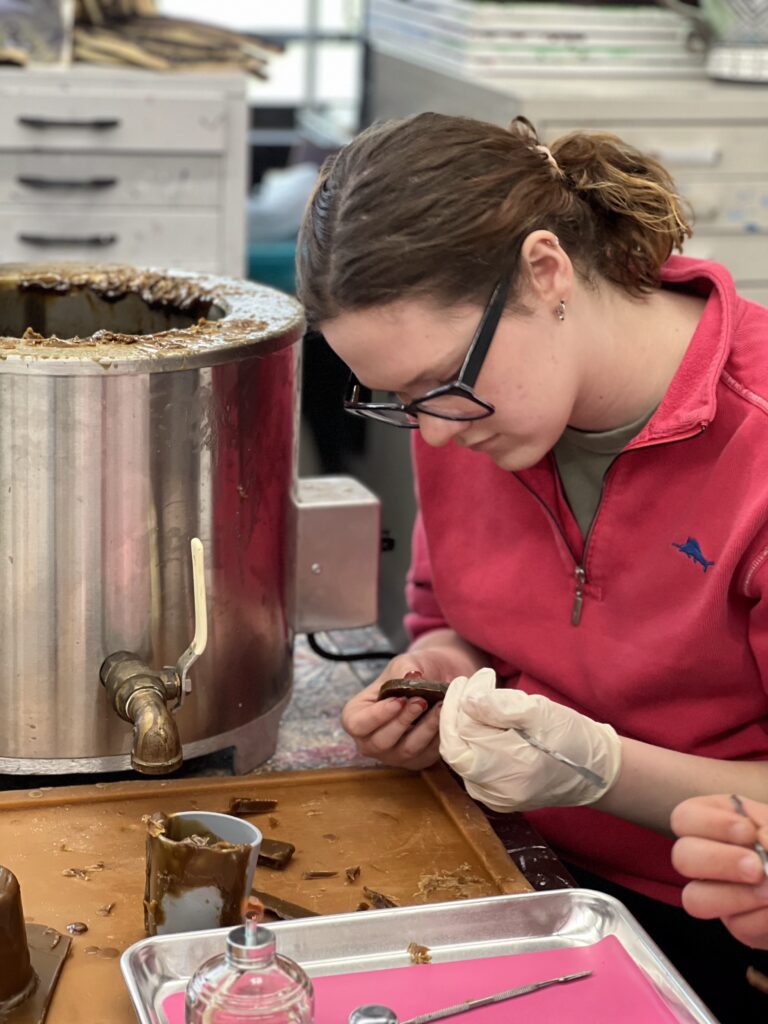 A person in a red jacket and glasses works on a belt buckle at a workshop, using a mold and tools. A large metal pot is nearby.
