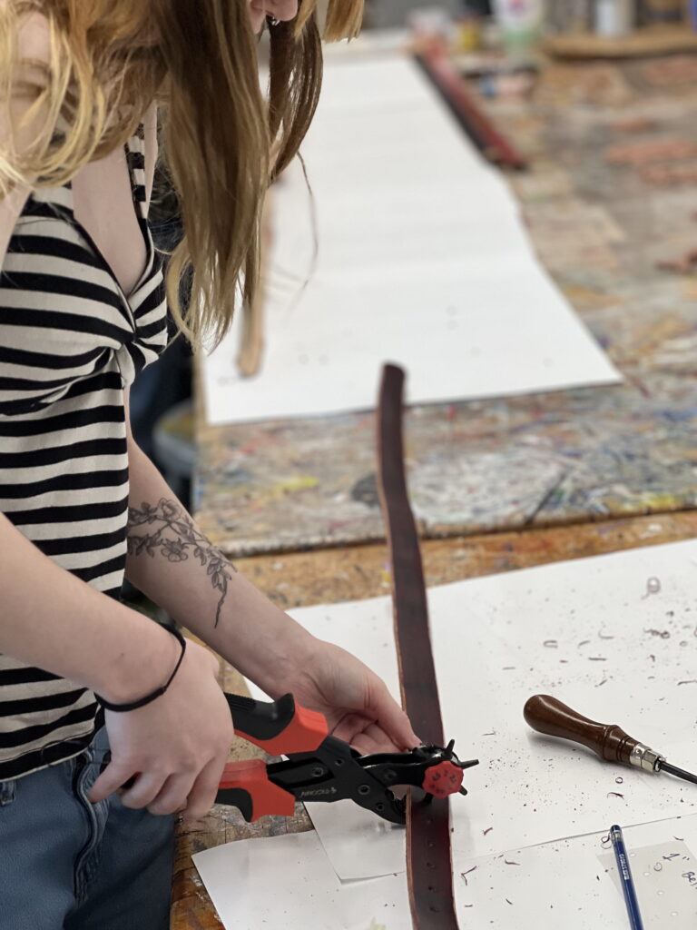 A person in a striped top uses a hole punch on a leather belt at a workshop table, with tools and paper scattered around.