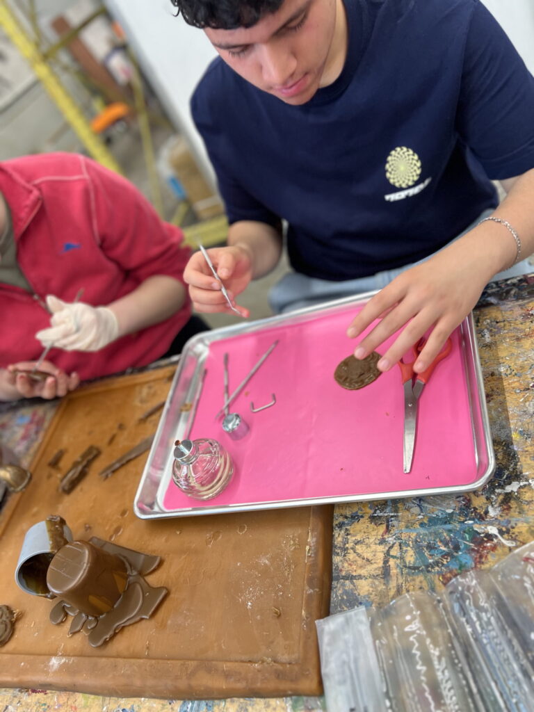 A person in a navy shirt works on crafting a belt buckle, using tools on a pink tray. Another person in red is partially visible.