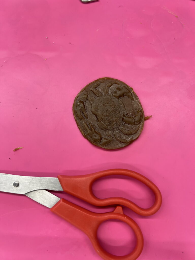 A pair of red-handled scissors next to a round, brown clay piece on a pink surface, likely part of a belt buckle workshop.