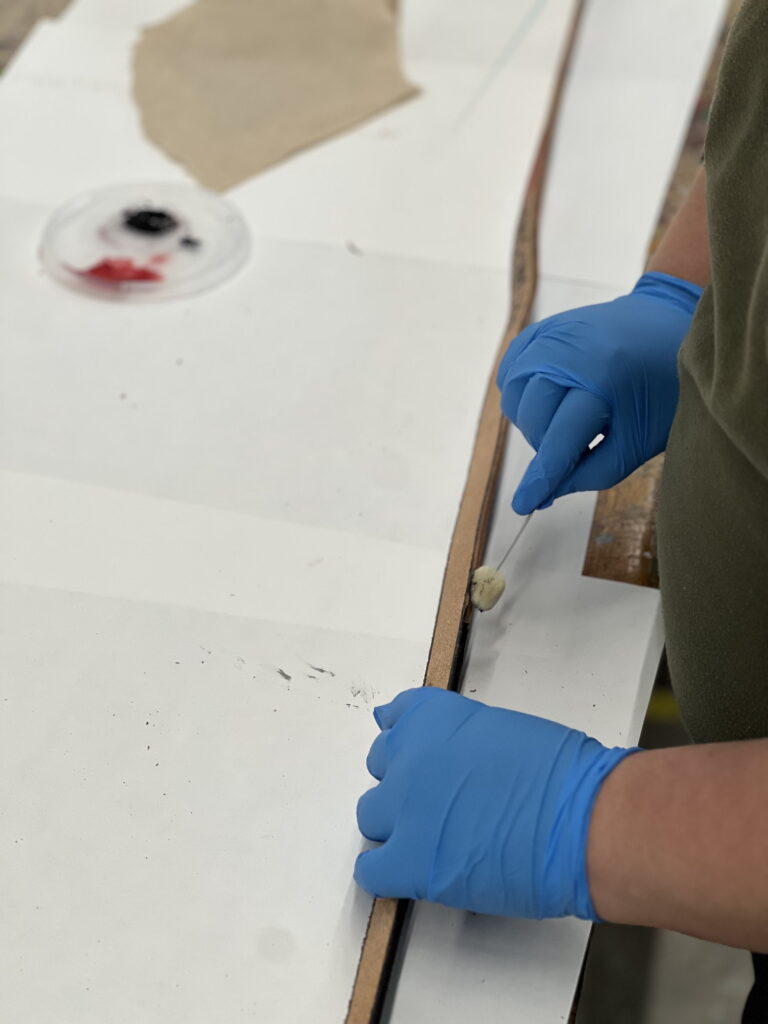 A person wearing blue gloves works on a project at a "make your own belt buckle workshop," applying a tool to a strip of material.