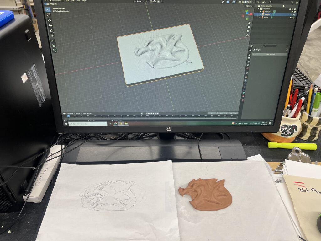 A computer monitor displays a 3D dragon design. Below, a sketch and a clay model of the dragon are on the desk.
